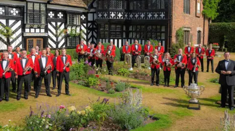 Members of the brass band, dressed in red jackets with the Foden's band logo on their lapels, are spread around the lawned areas of an ornamental garden in front of a black and white half-timber frame building. There is a large competition trophy in front of them.