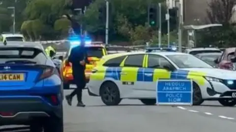 A police sign is in the middle of the road and says Heddlu. Police slow.  A police car is behind and a officer is pictured walking across the road