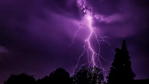 BroganJay87 Purple-coloured lightning strikes behind some trees during the night.