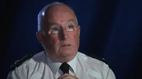 Spt Gerry Murray sitting before a blue background wearing a white shirt, PSNI eppolettes, and black tie and glasses