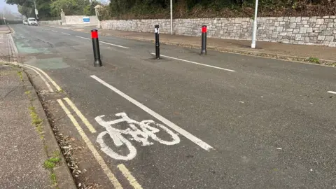 A white symbol of a bicycle painted on Dryden Road to show a cycle lane and bollards stopping motorised traffic from entering the road.