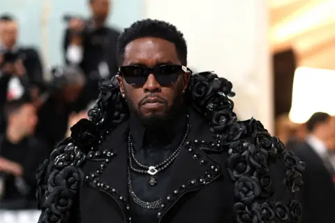 Reuters Sean Combs in an elaborate black outfit decorated with black roses and embellishments, surrounded by people in the background, some taking photos.