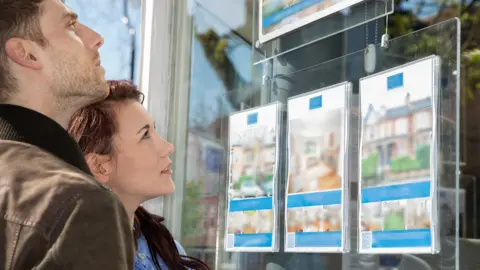 Close up of a young couple looking into an estate agents window showing both faces from the side and information on three houses in the window 