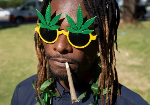 IHSAAN HAFFEJEE/REUTERS A man holds a cannabis cigarette in his mouth at the protest in Johannesburg. He is wearing yellow sunglasses with a big, green, marijuana leaf motif.