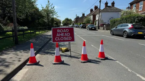 Badshot Lea Road near Farnham. A red sign says 'ROAD AHEAD CLOSED' in white letters. There are four red and white traffic cones in front of it. There are trees and plants on one side of the road, on the other are houses and cars.