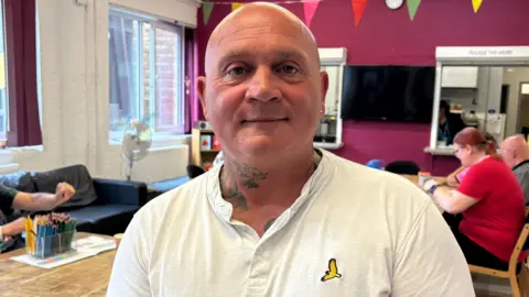 A man standing in a room, with people behind him. He is wearing a white T-shirt and has tattoos on his neck. He has a bald head and earrings. The wall behind him is purple.