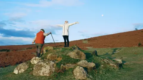 Frankie Mills Man in orange gilet with walking stick walks towards woman standing on rock with eyes closed and arms outstretched
