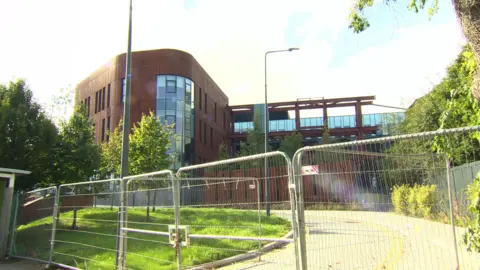 Large red clad building in the background- path with grass and trees in front all blocked off by tall fencing 