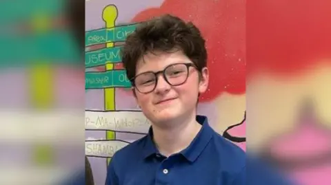 Supplied A teenage boy wearing glasses with brown hair smiles at the camera. He is standing in front of a wall painted with colourful artwork.
