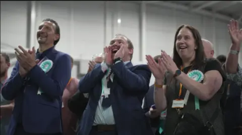 Three Reform candidates or supporters stand together and applaud at the May election result night. Two men are wearing navy suits and a woman is in a brown short-sleeved top. All three have green and white party rosettes on.
