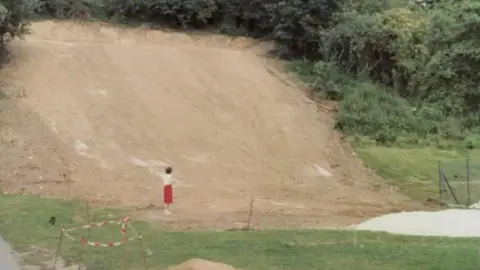 Kendal Snowsports Club A grainy image showing a woman wearing a red skirt and a white top who stands beneath a steep earthy slope. The slope has green grass on the side and at the bottom. There is red and white tape marking off parts of it.