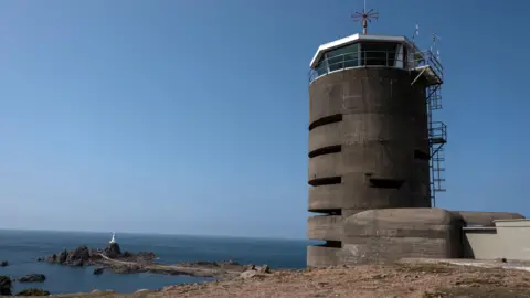 A brown concrete radio tower stands out on he coastline next to the blue of sea and sky