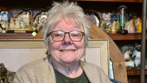 Jayne smiles into the camera. She is sat in her house surrounded by the items she collects, including ornamental cats and framed pictures. She has glasses, short grey hair and is wearing a light brown cardigan over a green patterned top.