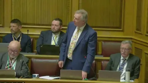 A man in a blue blazer and a yellow tie, standing up to speak in a panelled room. There are four other suited men sitting around him.