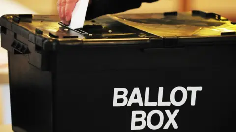 A person casts their vote in a black ballot box with ballot box in white letters on its side.