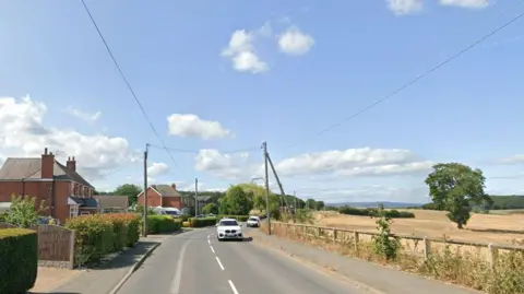 A road whish has houses on one side and fields on the other, it is a summer day and the sky is blue. The fields look like they contain corn and there are two white cars on the road. 