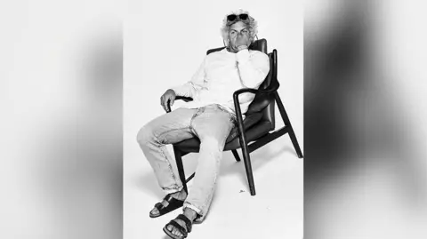 Dan Landsburgh Tom Jackson in a black and white shot. He is lounging in a black leather chair in front of a white background. He's wearing jeans, no socks and Birkenstock sandals with a white cotton long sleeved round neck top. He has his hand up in a fist in front of the lower half of his face. He had long curly black and white hair and has a pair of dark sunglasses perched on his head.