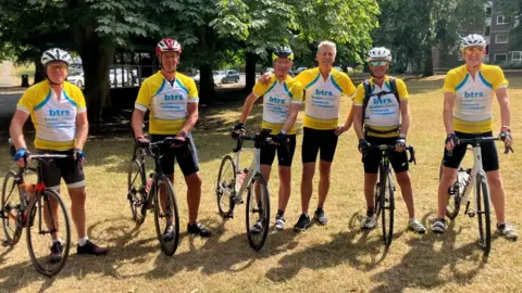 Six men wearing yellow branded cycling jerseys which read "B T R S, brain tumour research and support". They are standing with bicycles in a park, and most wear cycling helmets.