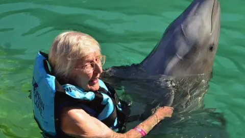 Family handout  Pauline Polhill swimming with a dolphin. She is an elderly lady with fair hair and large glasses. She is wearing a blue lifejacket and looking to the side of the camera. The dolphin is in front of her, with its head sticking up out of the water.