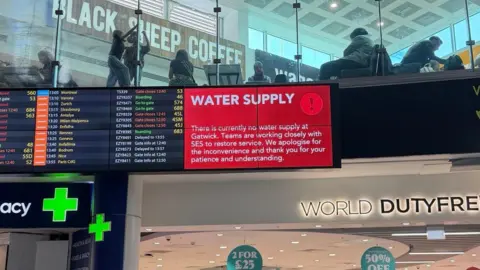 Supplied A red sign alerts passengers to the water supply issue on a screen in a terminal building. A World Duty Free sign is underneath and a pharmacy sign is nearby. A further screen shows flight information.