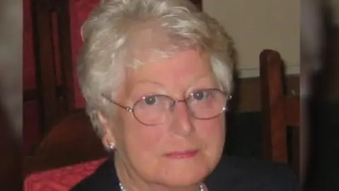 Janet Purkess is seated in a room. She has neat, wavy, short, silver hair and glasses.