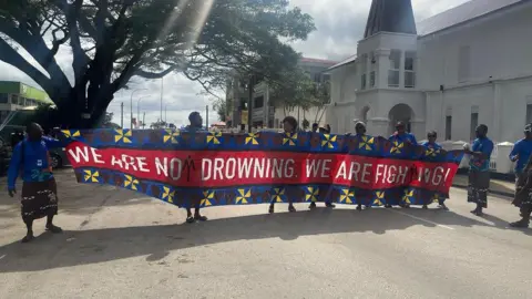 BBC A women's group holds a banner in the streets that reads 'We don't drown, we fight'