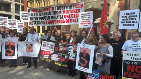 A large group of people holding placards outside the Lampard Inquiry in London. Their placards criticise mental health trusts in Essex and call for action.