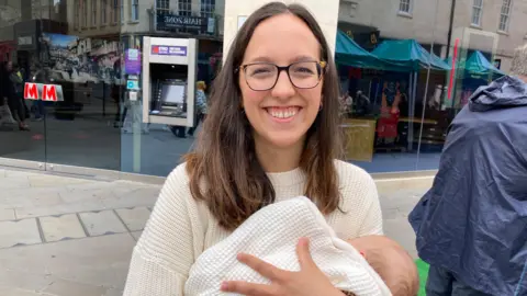 Carmen Murillo stands in the street holding her three-month-old daughter, who is wrapped in a white blanket. Carmen has medium-length brown hair, wears thin glasses and is smiling. She is wearing a knitted cream-colored sweater.