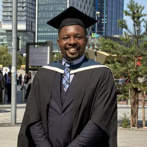 A man wearing a dark blue suit and tie with a black gown and mortar board over the top. Behind him are trees and tall glass buildings with apartments and offices.