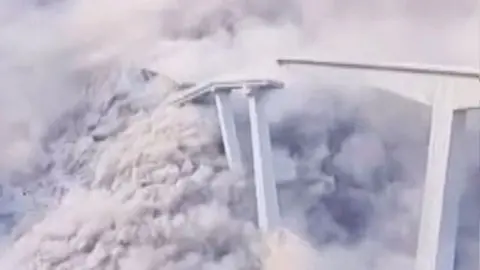 Bridge collapses leaving clouds of white smoke and dust, part of the remaining bridge is on the right of the image while the collapsed part of the bridge is on the left of frame.