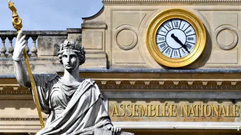 Getty Images The French National Assembly is seen in this close-up photograph, framing the sign and golden clock on the facade and also the statue of a woman holding a golden sceptre which stands before the entrace