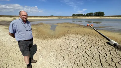 Mike Liggins/BBC Tony Bambridge, a Norfolk farmer, in front of a largely dried up reservoir