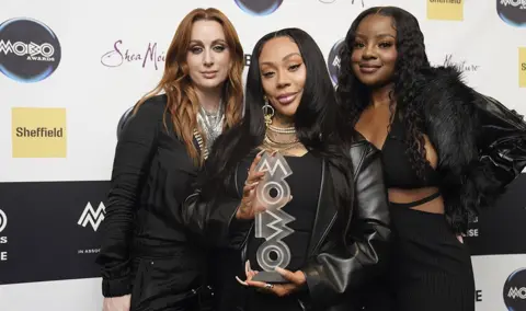 PA Media The Sugababes pictured holding their Impact Award in front of a white billboard with Mobo logos and advertising