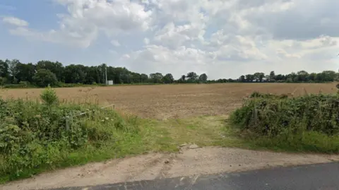 Google A field of bare soil is shown with a low hedgerow and an open metal gate in the foreground of the image and with trees bordering it in the background.