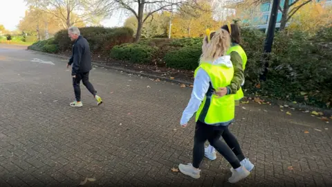 Gary Shaughnessy on the left walking through a car park with Katie Tyler and Sophie Drew, who are tied together and wearing high visibility vests and Pudsey ears.