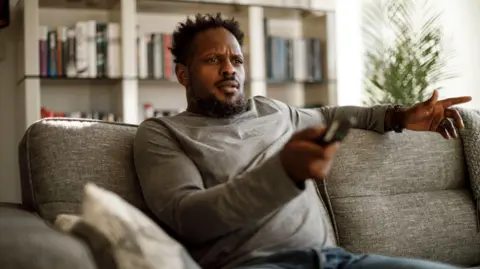 Getty Images Man sitting connected  a sofa holding a TV distant    with a confused expression