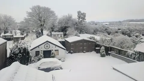Weather Watcher Sarah The back garden of house is covered in snow. There are tow small sheds. The one of the left is white, with a snow covered roof and the one on the right is black with a snow covered roof. A car sits in front of the white shed, covered in snow. Behind the garden is a black wooden fence. Beyond the fence is another garden and a group of tall trees covered in snow. 