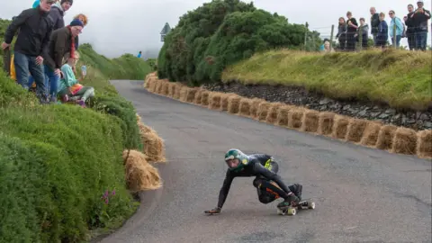 WDSC A skateboarder in black leaning over coming down on a skateboard. Hay bales line the road and crowds are watching from the hedgerows on either side.