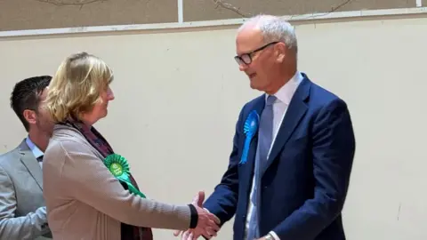 Louise Parker/BBC A man dressed in a blue suit is shaking the hand of a woman wearing a grey jumper with a green party rosette. 