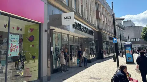 A photo of a high street with Marks and Spencers. Dozens of people are walking along the street, with Primark in the background