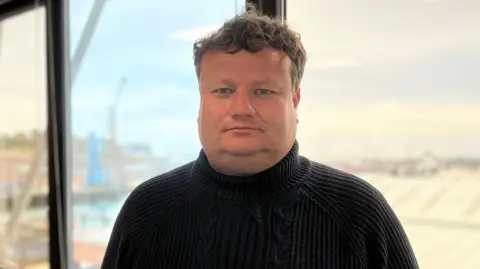 William is in a dark sweater standing indoors near a large window overlooking a harbour area.