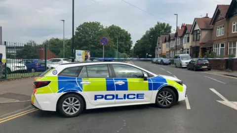 A police car that is parked in front of a blue police cordon on a residential road. There are terraced houses on the right side of the road and a school on the left side behind a green fence. 