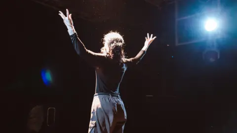 A performer on stage dressed in a black top and grey trousers. 
A spotlight can be seen at the side of the stage.