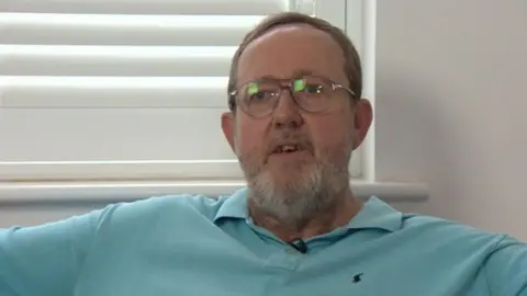 A man in a light blue polo shirt sits in a white-walled room with white shutter blinds on a window behind him. He has short dark hair and a white/brown beard. He wears metal-rimmed glasses and his mouth is slightly open as if about to speak.