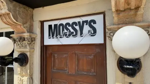 A sign depicting Mossy's Nightclub above a door, with decorative masonry and white glass globe lamps on black ironwork fittings.