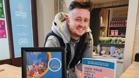 A man with brown spiky hair that is shavedd at the sides is wearing a grey hoody and black gilet while leaning on the counter of shop. In front of hime is a framed sign and a book with the words feed your family for a 