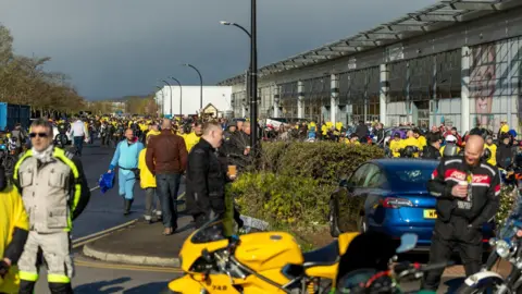 Roadrunnerpics A large outdoor gathering of motorcyclists in what appears to be a car park. The area is busy with people, many dressed in riding gear. Several participants are wearing bright yellow outfits.