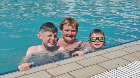 Family handout Two young boys and a woman are in a swimming pool. The child on the right side of the image is wearing swimming goggles. The woman in the middle has blond hair. The two boys both have dark hair. 