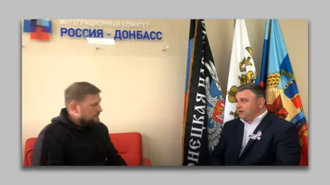 Southern Front The logo of the Russia-Donbas Integration Committee visible on the wall above two men conducting an interview