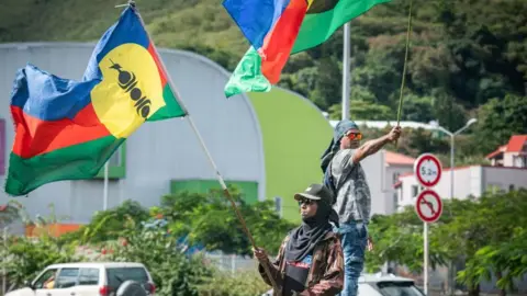 Getty Images Pro-independence supporters wave flags on the side of a road ahead of legislative elections in the Vallee du Tir district in Nouméa on the French Pacific territory of New Caledonia on June 27, 2024. 